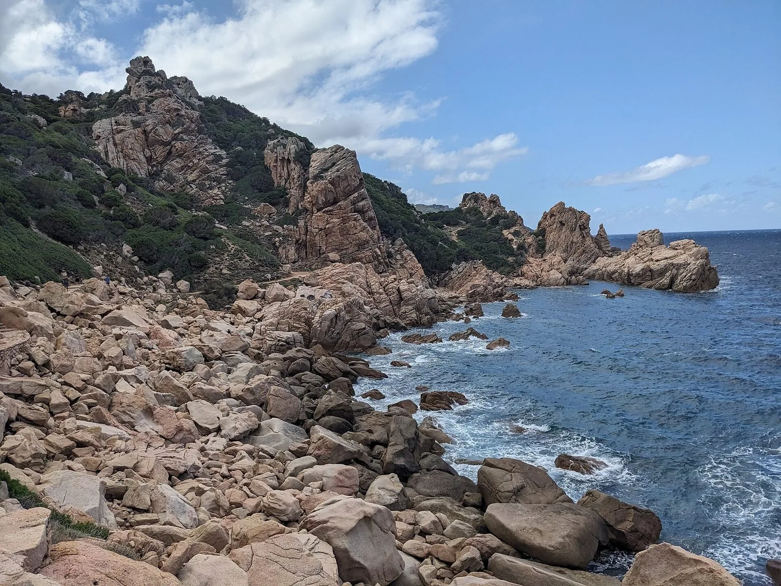Scogliera di roccia rosata e massi lungo il sentiero per Li Cossi a Costa Paradiso, con il mare blu increspato